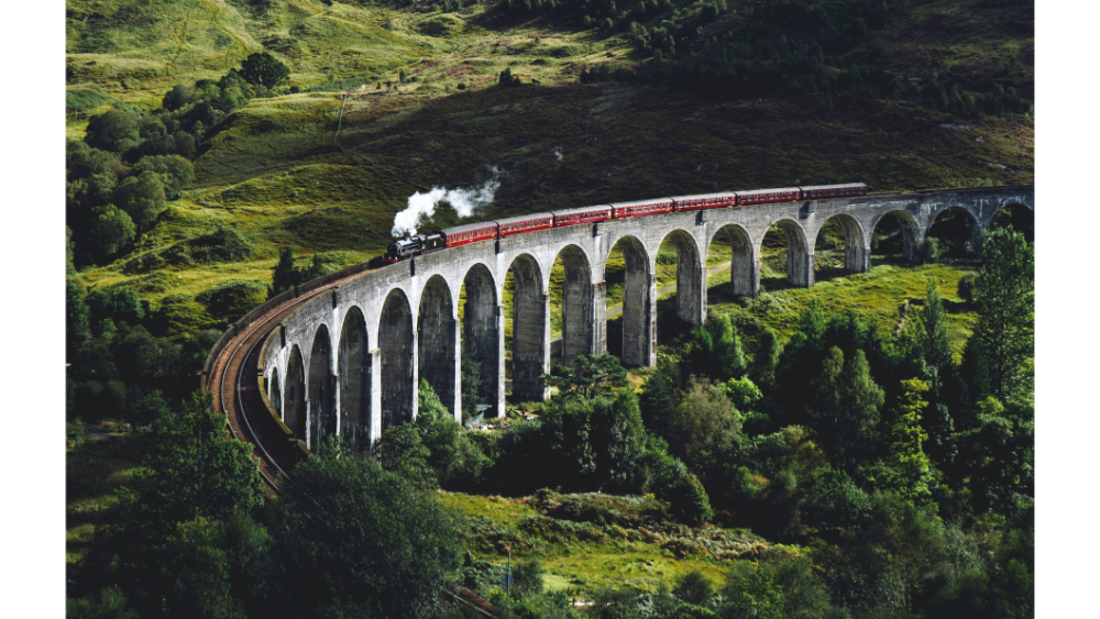 Steam train with red carriages crossing a tall stone viaduct in a lush green valley.