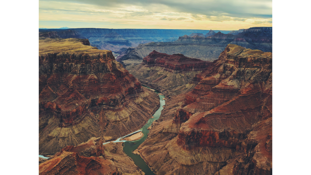 Expansive view of the Grand Canyon with layered red rock formations and the Colorado River winding through the canyon floor under a cloudy sky.