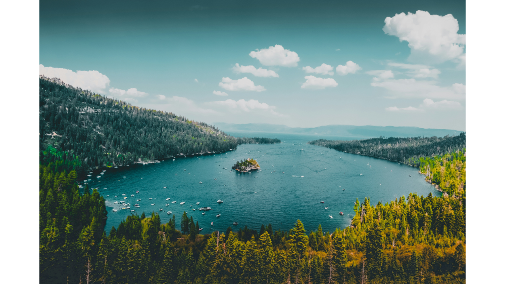 Aerial view of Lake Tahoe surrounded by dense pine forest and distant mountain ranges under a bright blue sky.