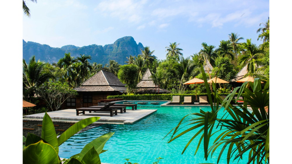 Tropical resort in Thailand with a turquoise swimming pool, palm trees, sun loungers, and wooden villas surrounded by lush green mountains.
