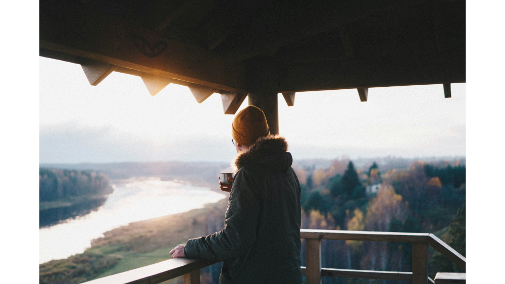Person in a winter coat and beanie enjoying a cup of coffee while overlooking a scenic river valley from a wooden balcony at sunrise.