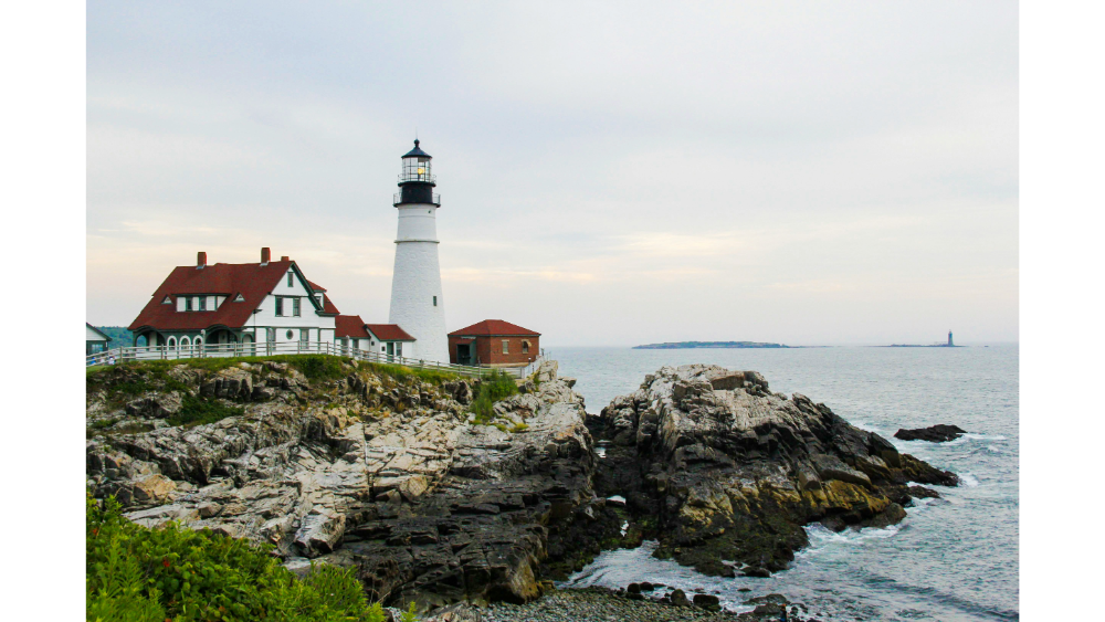 Coastal lighthouse and buildings on rocky shoreline overlooking the ocean on a cloudy day.