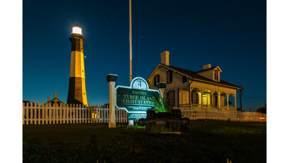 Night view of the historic Tybee Island Lighthouse with its adjacent keeper's house illuminated under a starry sky.