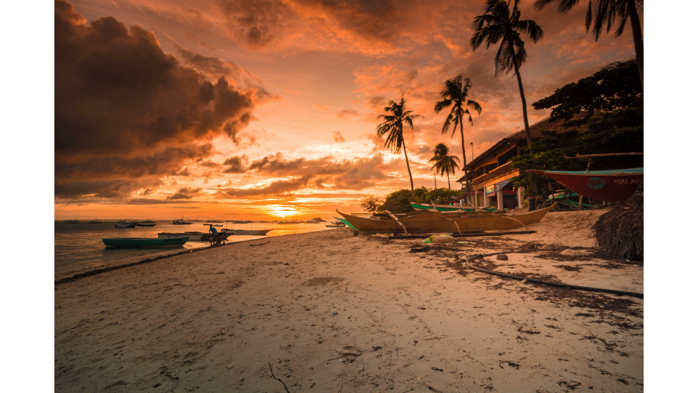 Tropical beach at sunset with palm trees, traditional boats on the sand, and dramatic clouds over a glowing horizon.