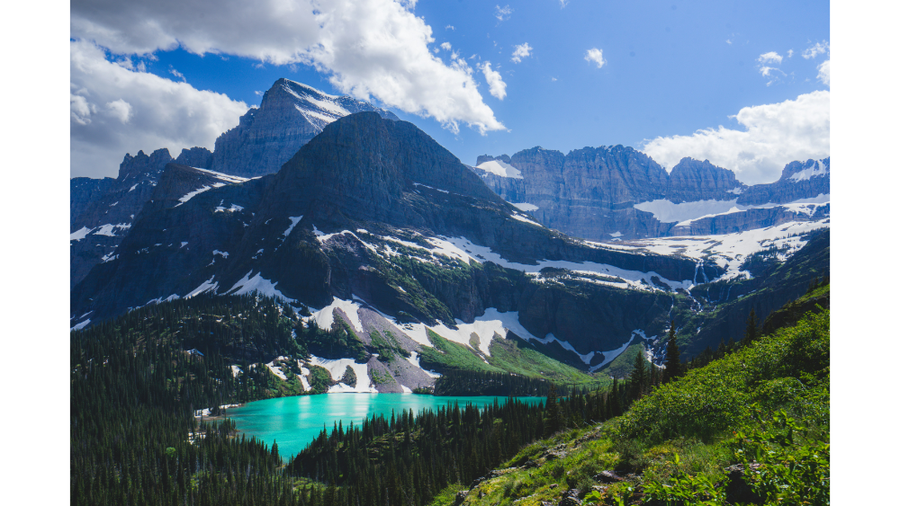 Turquoise alpine lake surrounded by snow-capped mountains and pine trees in Glacier National Park under a bright sky.