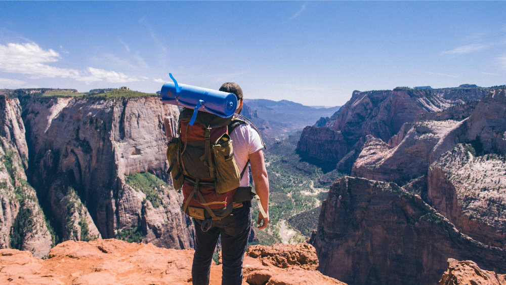 Solo backpacker standing on a rocky cliff edge with a rolled-up sleeping pad, gazing across a vast canyon landscape.