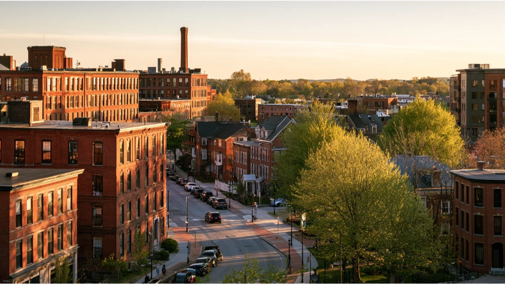 Commuter vehicles traveling along a Manchester, New Hampshire, roadway near local businesses and traditional New England buildings on a typical weekday.