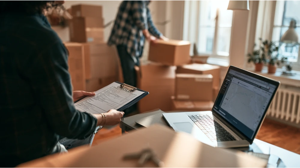 Couple unpacking in an apartment with moving boxes, a rental checklist, and a laptop used for home searching.