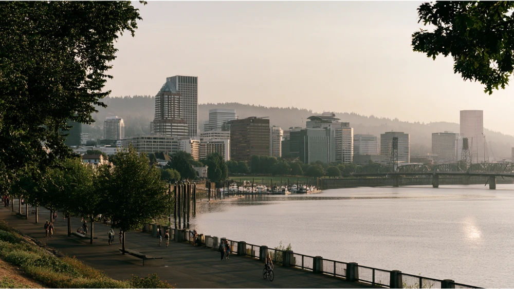 Portland, Oregon, skyline along the Willamette River with downtown buildings, green riverfront parks, and the surrounding Pacific Northwest landscape.