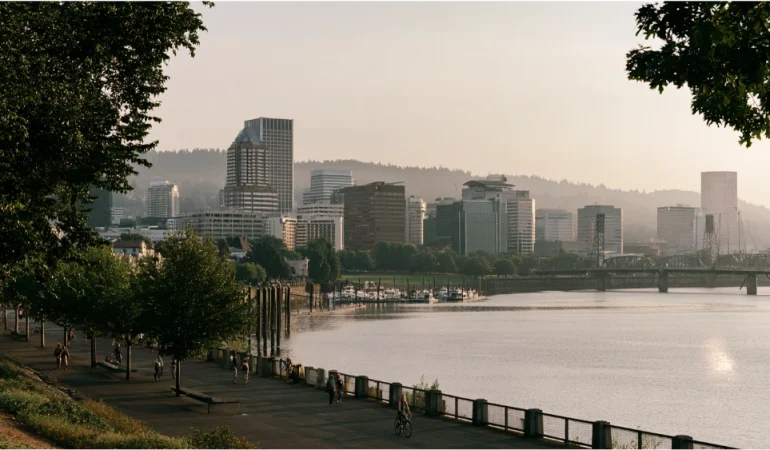 Portland, Oregon, skyline along the Willamette River with downtown buildings, green riverfront parks, and the surrounding Pacific Northwest landscape.